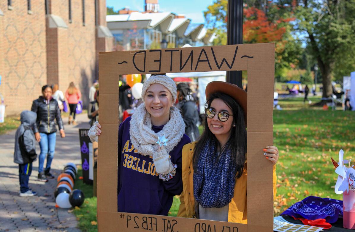 Two female students hold a wanted poster frame around their faces while smiling for the camera during the Octagon Fair